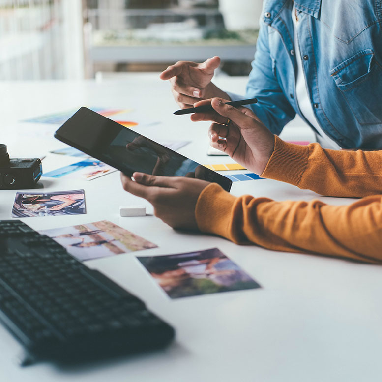 Zwei Personen an einem Schreibtisch, eine davon mit Tablet und Stift. Verschiedene Fotos, Farbmuster und eine Kamera sind auf der Oberfläche verteilt. Im Vordergrund ist eine Tastatur zu sehen, die auf einen Kreativ- oder Designarbeitsplatz hindeutet.