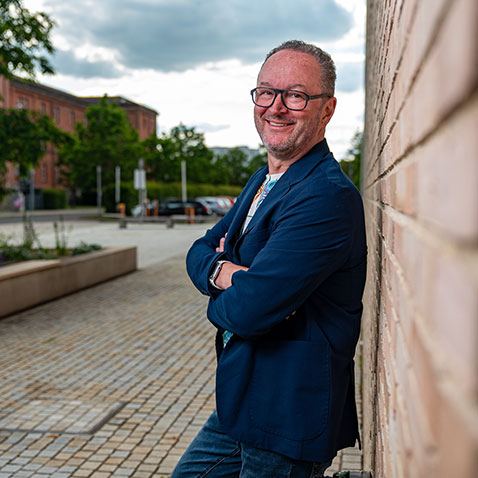 René Reiter Werbeagentur Eine Person mit Brille und Bart steht im Freien an einer Backsteinmauer. Sie lächelt und hat die Arme verschränkt. Im Hintergrund sind ein gepflasterter Gehweg, Gebäude mit Grünflächen und ein bewölkter Himmel zu sehen.