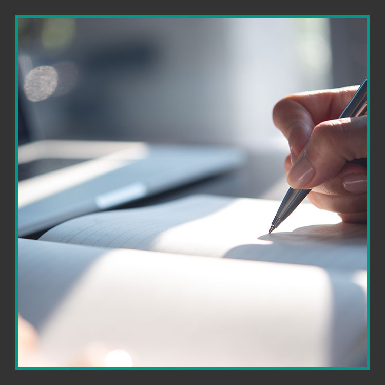 Schreiben Datenschautz Digitalagentur Werbeagentur Ansbach Nahaufnahme einer Hand, die einen Stift hält und in ein Notizbuch schreibt. Die Szene ist gut beleuchtet, Sonnenlicht wirft weiche Schatten auf die Seiten. Im Hintergrund sind ein unscharfer Laptop und ein weiteres Notizbuch zu sehen.