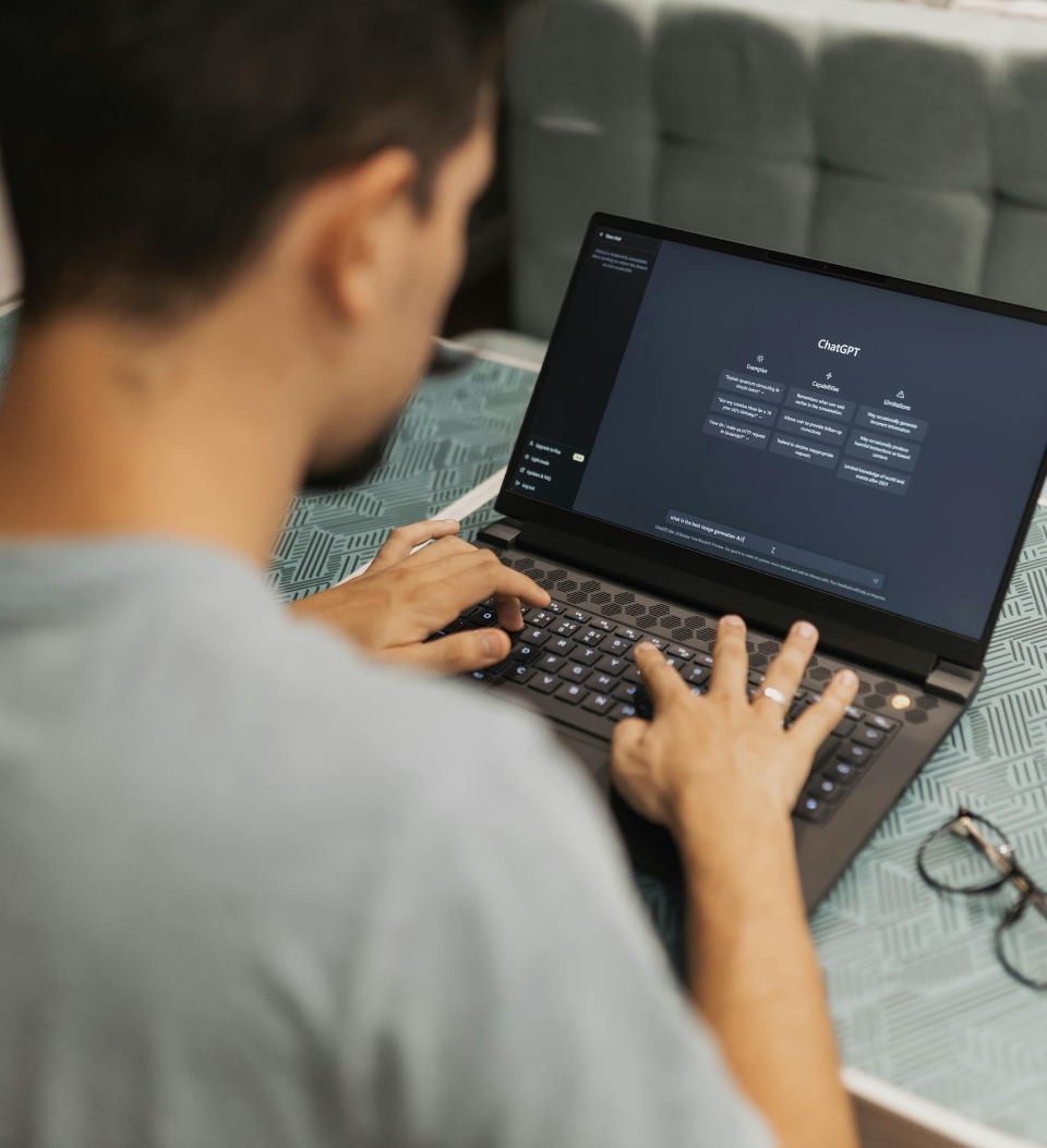 Eine Person tippt auf einem Laptop, der die ChatGPT-Schnittstelle anzeigt. Der Laptop steht auf einem gemusterten Tisch, daneben liegt eine Brille. Das Bild ist auf den Laptop und die Hände fokussiert, wobei das Gesicht der Person nicht zu sehen ist.