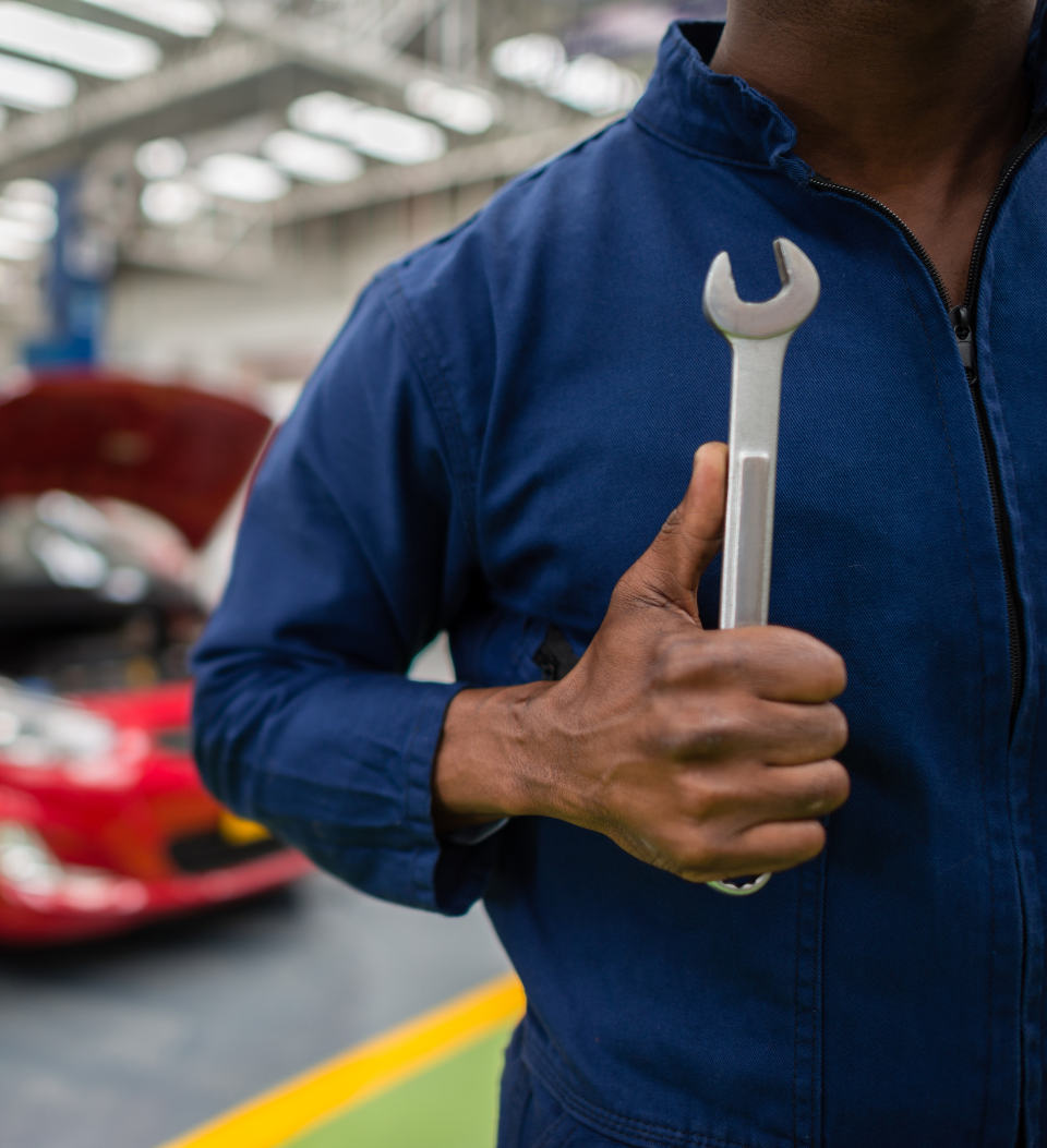 Ein Mechaniker in einem blauen Overall hält in einer Autowerkstatt einen großen Maulschlüssel gegen die Brust, während im unscharfen Hintergrund ein rotes Auto und andere Werkstattausrüstung zu sehen ist.